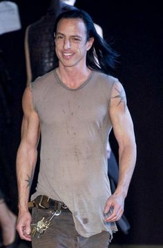 a man with long black hair walking down a runway wearing a tank top and jeans