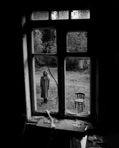 a black and white photo of a person standing in front of a window with a chair