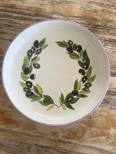 a white bowl with an olive wreath painted on the side sitting on a wooden table