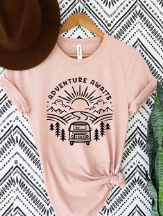 a t - shirt with the words adventure await on it next to a hat and plant