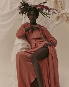 a woman in an orange dress sitting on a chair with her legs crossed and wearing a flower crown over her head