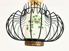 a white vase with green leaves on it is hanging from a black wire holder that has gold trim around the neck