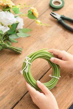 a person holding a green wreath on top of a wooden table next to scissors and flowers