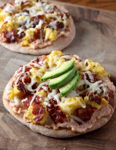 two small pizzas with cheese, bacon and an avocado on them sitting on a cutting board