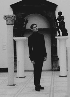 black and white photograph of man standing in front of statues on pedestals, looking up at the sky