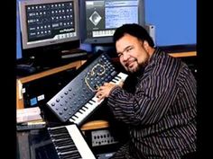 a man sitting in front of a keyboard with multiple electronic equipment around him and smiling at the camera