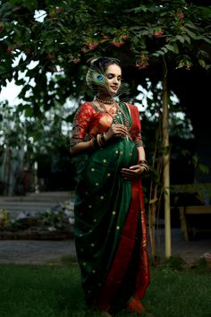 a woman in a green and red sari standing under a tree with her hands on her hip