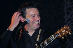 a man holding a guitar up to his ear while singing into a microphone and smiling