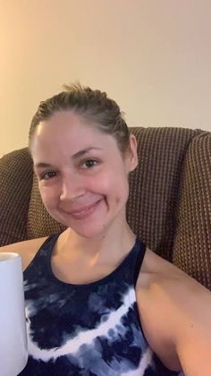 a woman sitting on a couch holding a white coffee mug in her right hand and smiling at the camera