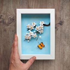 a hand is holding a shadow box with a butterfly on it and flowers in the background