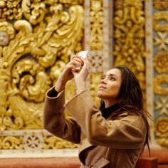 a woman taking a photo with her cell phone in front of ornate gold wallpaper