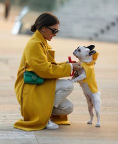 a woman kneeling down petting a small dog wearing a yellow coat and white pants