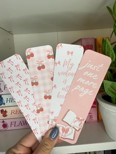 a hand holding four pink and white bookmarks in front of a shelf with books