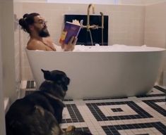 a man sitting in a bathtub reading a book next to a dog on the floor