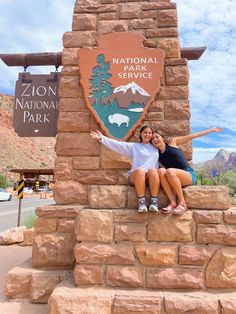 two women are posing for a photo in front of the national park sign and mountains