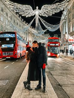 a man and woman standing on the sidewalk in front of double decker buses at night