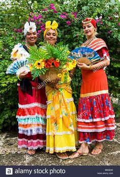 three women in colorful dresses holding flowers and fan standing next to each other on the ground