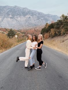 three women standing in the middle of an empty road with mountains in the back ground