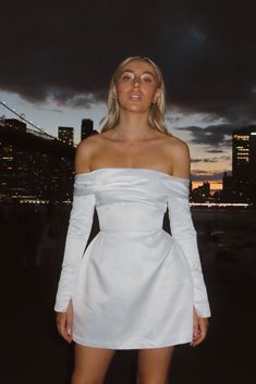 a woman standing in front of a cityscape at night wearing a white dress