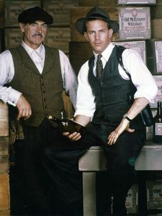 two men in vests and hats sitting on a bench