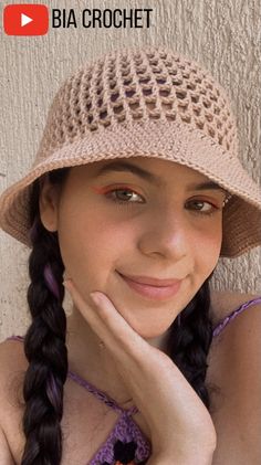 a girl wearing a hat and smiling with her hand on her chin, in front of a wall