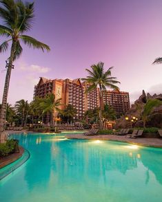 an outdoor swimming pool surrounded by palm trees and hotels in the background at dusk or dawn