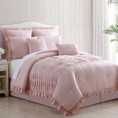 a bed with pink comforter and pillows in a room next to a plant on a table