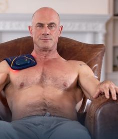 a bald man with no shirt on sitting in a chair holding an electronic device to his chest