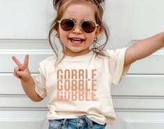 Celebrate Thanksgiving in style with our adorable Gobble Gobble t-shirt for girls! This festive and fun shirt is perfect for your little one to wear on Turkey Day, whether she's helping in the kitchen, enjoying the family feast, or participating in Thanksgiving activities. With a cute turkey and playful "Gobble Gobble" text, your little turkey will be the center of attention! Product Details: Design: "Gobble Gobble" in playful, trendy lettering Material: Soft, breathable cotton blend for a comfortable all-day fit Sizes: Available in toddler, baby and youth sizes. Fit: Classic fit, perfect for layering with cardigans or jackets Care Instructions: Machine wash cold, tumble dry low Why You'll Love It: -Adorable Thanksgiving-themed design with fun "Gobble Gobble" text -Soft and cozy fabric per Funny Toddler Thanksgiving Shirt, Diy Thanksgiving Shirts For Kids, Thanksgiving Shirts For Kids, Kids Thanksgiving Shirts, Trendy Lettering, Kids Shirts Design, Cute Turkey, Funny Thanksgiving Shirts, Turkey Shirts