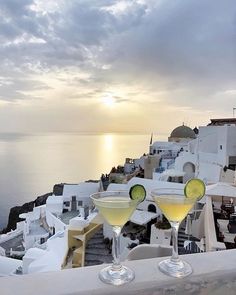 two martini glasses with cucumber slices sit on a ledge overlooking the ocean and white buildings