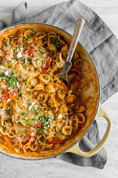 a skillet filled with pasta and vegetables on top of a cloth next to a wooden spoon