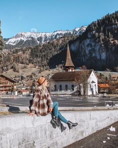 a woman sitting on top of a cement wall next to a mountain town with snow capped mountains in the background