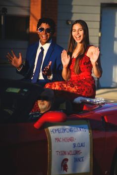 a man and woman sitting on top of a red car waving to the camera with their hands in the air