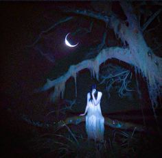 a woman in white dress standing under a tree at night with the moon behind her