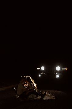 a woman sitting on the ground in front of a car at night with headlights shining