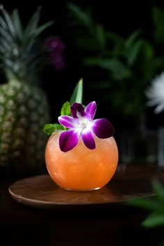 an orange vase with purple flowers in it on a wooden plate next to pineapples