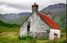 an old building with a red roof in the middle of a grassy field next to mountains