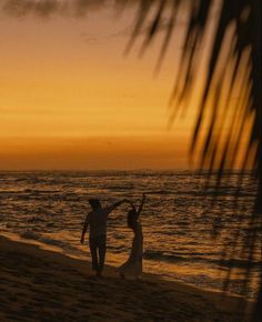 a man and woman walking on the beach at sunset with their arms in the air