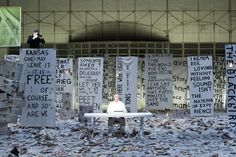 a man sitting at a white table surrounded by protest signs in front of a building