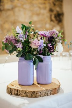 two purple mason jars with flowers in them sitting on a wood slice at a wedding reception