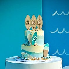 a three tiered cake decorated with surfboards and the word one on top is sitting on a table in front of a blue wall