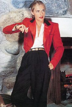 a woman in a red jacket and black pants standing next to a fire place with her hands on her hips