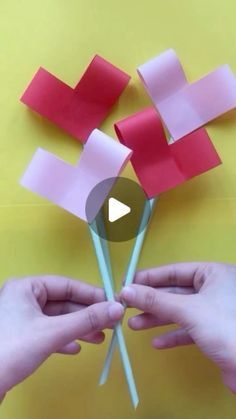 two hands holding red and pink paper flowers with sticks sticking out of them on a yellow background