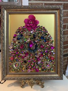 a framed painting with a pink flower and jewels in it on a table next to a brick wall