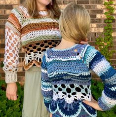 two women standing next to each other in front of a brick wall wearing crocheted sweaters