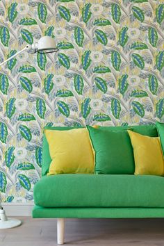 a green couch with yellow pillows in front of a wallpapered background and floor lamp