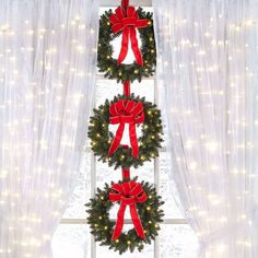 three wreaths with red bows are hanging on the window sill in front of white curtains