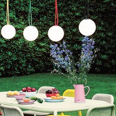 an outdoor table and chairs with fruit on the table in front of some bushes, hanging lights and flowers