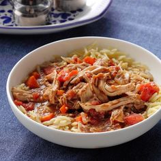 a white bowl filled with pasta and meat on top of a blue table cloth next to silverware