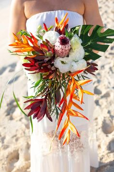 a woman in white dress holding a bouquet of flowers on the beach with palm leaves